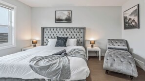 A bedroom with a large bed, two bedside tables, a chair, and two wall-mounted pictures. - Luxurious  4-bedroom house w/ Free Banff Pass (Northeast)