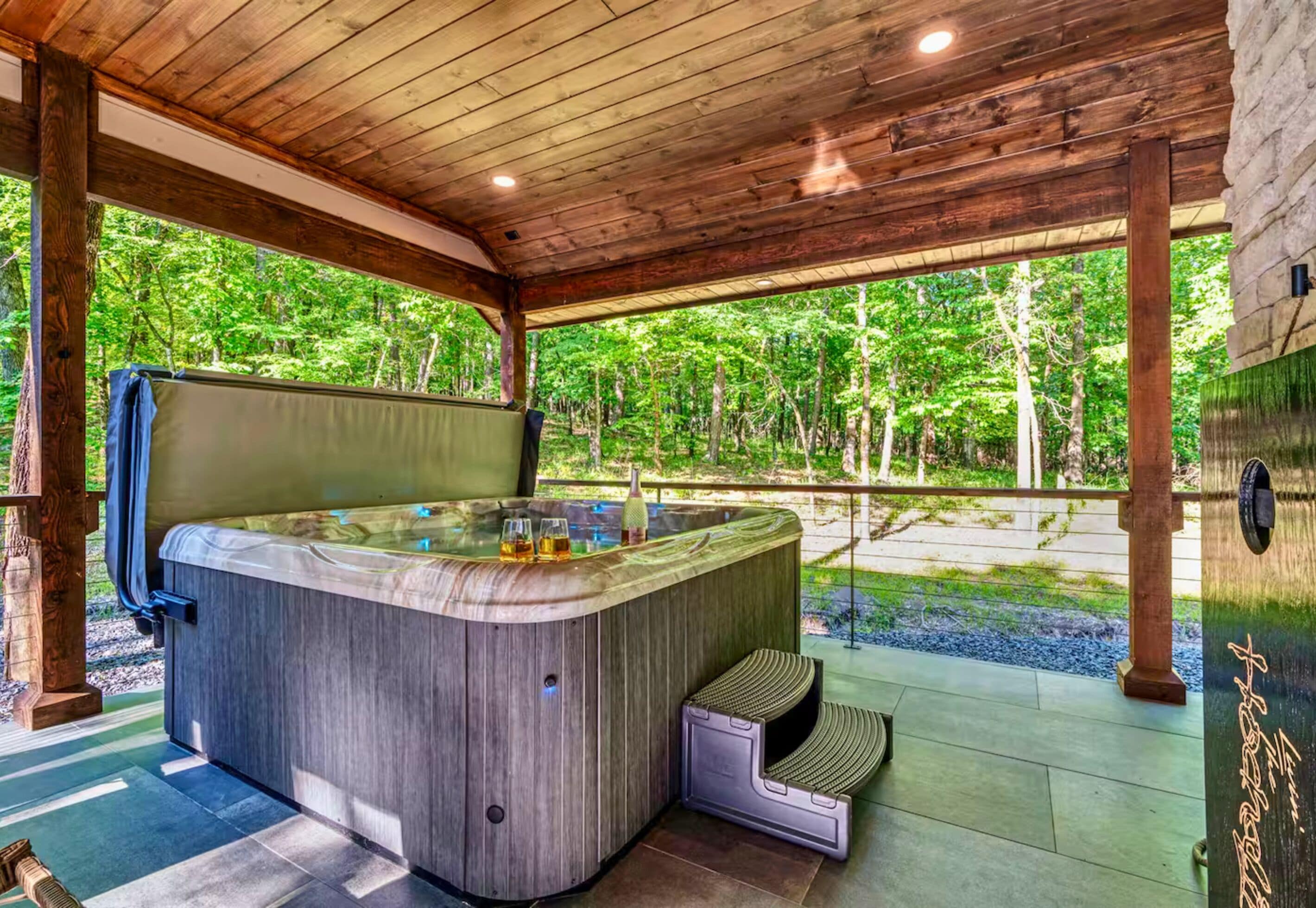 Ein Whirlpool mit Holzaußenverkleidung, Glasumrandung und einer Holzterrasse mit Blick auf einen Wald.