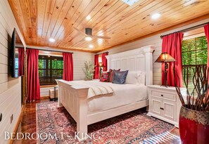 A bedroom with a wooden ceiling, a bed with a white headboard, a nightstand, a lamp, and a window with red curtains. - Heated Pool | Boat Parking | Outdoor Kitchen (Broken Bow)