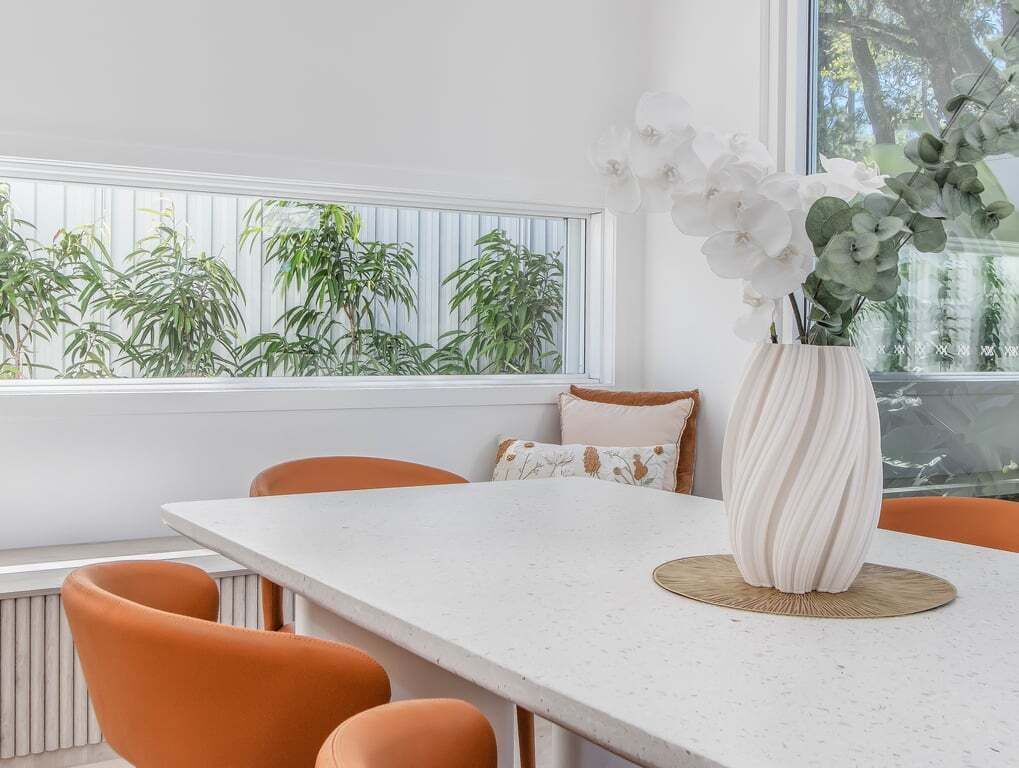 Un comedor con una mesa blanca, sillas anaranjadas, un jarrón con flores blancas y una ventana con plantas.