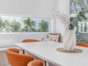 A dining area with a white table, orange chairs, a vase with white flowers, and a window with plants. - 23 Lancelin Drive Mermaid Waters (Gold Coast)