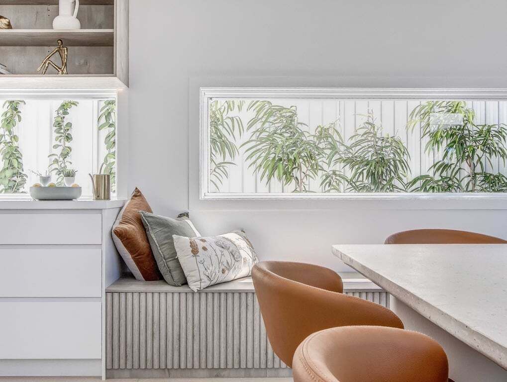 A room with a white radiator, a bench with cushions, a table, and a window with a view of plants.