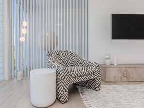 A modern living room with a patterned armchair, a white ottoman, a wooden cabinet, and a large window with blinds. - 23 Lancelin Drive Mermaid Waters (Gold Coast)
