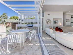 A modern outdoor patio with a dining table and chairs, a white sofa, and a kitchen area visible through an open door. - 23 Lancelin Drive Mermaid Waters (Gold Coast)