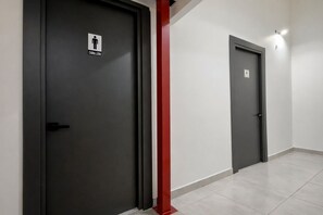Dos puertas de color oscuro con el símbolo de un baño de hombres, un pilar estructural rojo y un suelo de baldosas.