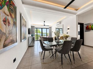 A modern dining area with a glass table and grey chairs, a ceiling fan, and a view to the outside. - Cozy 1-bedroom apartment in tranquil Sosúa with AC, Hot Water, Pool and GYM (Sosúa)