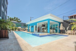 Un espace moderne aménagé au bord de la piscine, avec une piscine à l’eau d’un bleu limpide, un espace détente couvert et un bâtiment à la façade bleue.
