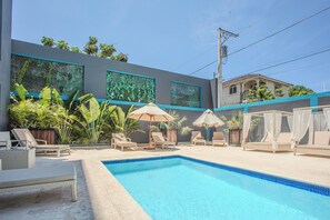 Piscina con sillones de descanso y sombrillas, rodeada de plantas tropicales y un edificio moderno con amplios ventanales.