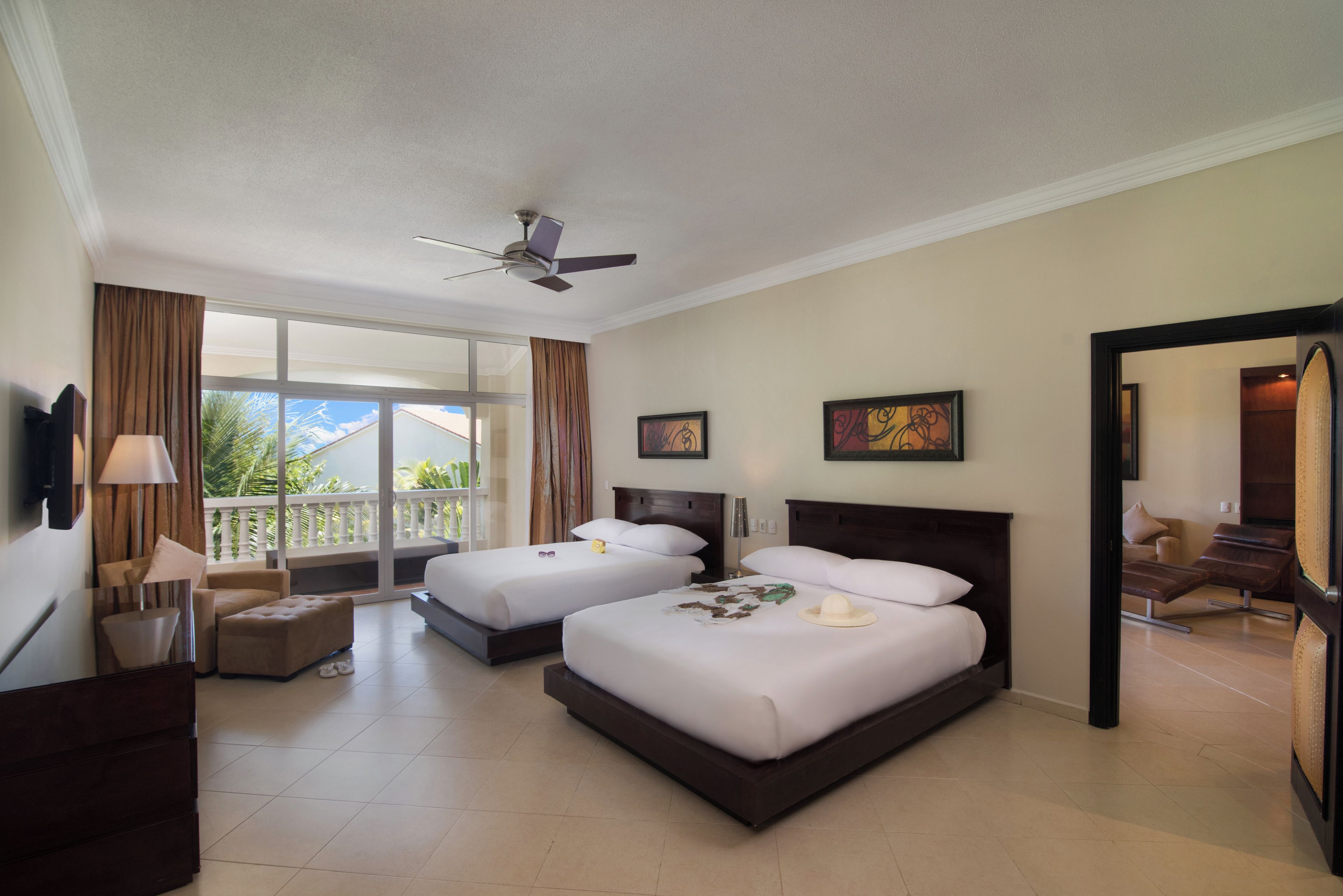 A hotel room with two beds, a ceiling fan, a balcony with curtains, and a view of the outdoors.