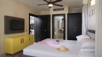 A hotel room with a bed, a television, a ceiling fan, a yellow cabinet, and a bathroom visible through a glass door.