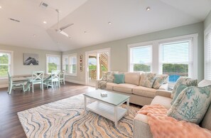 A spacious living room with a sofa, coffee table, and chairs. There are large windows with blinds, a ceiling fan, and a view of the outdoors. - The Fourth Watch - 4x4 Carova Beach, Ocean View, Brand New, Linens, Flex Stays (Carova Beach)