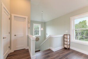 A hallway with a wooden floor, a white door, a window with a view of trees, and a small wooden shelf.