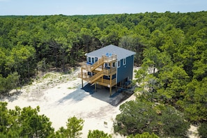 A two-story wooden structure with a deck and outdoor seating, surrounded by dense forest. - The Fourth Watch - 4x4 Carova Beach, Ocean View, Brand New, Linens, Flex Stays (Carova Beach)