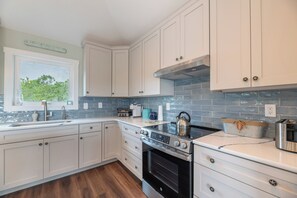 A modern kitchen with white cabinetry, a stainless steel range, and a window with a view of greenery. - The Fourth Watch - 4x4 Carova Beach, Ocean View, Brand New, Linens, Flex Stays (Carova Beach)