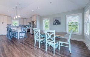 A spacious kitchen with modern appliances, a dining area with light blue chairs, and large windows with blinds. - The Fourth Watch - 4x4 Carova Beach, Ocean View, Brand New, Linens, Flex Stays (Carova Beach)