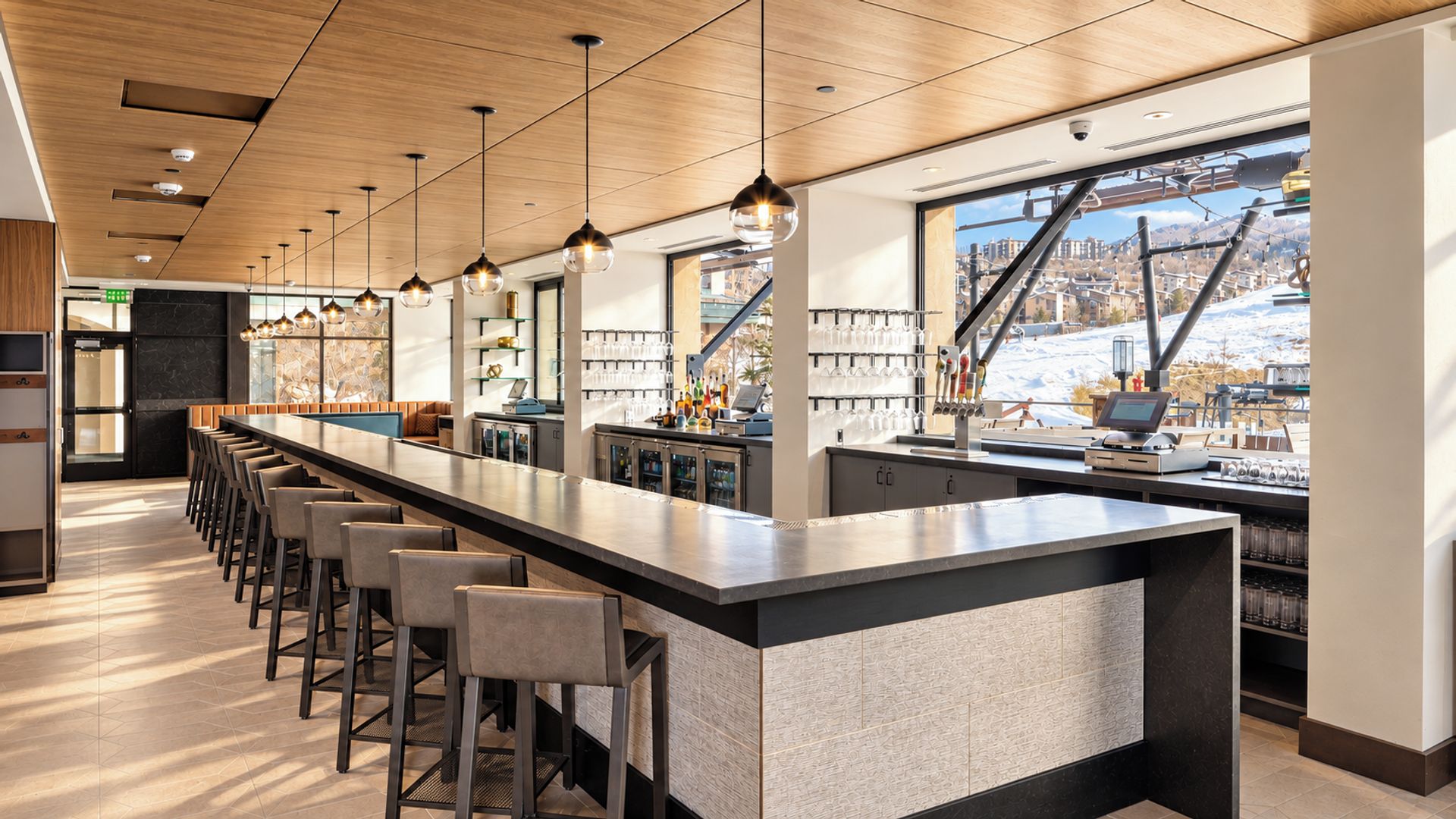 A modern bar area with a long counter, bar stools, pendant lighting, and a view of a snowy landscape through large windows.