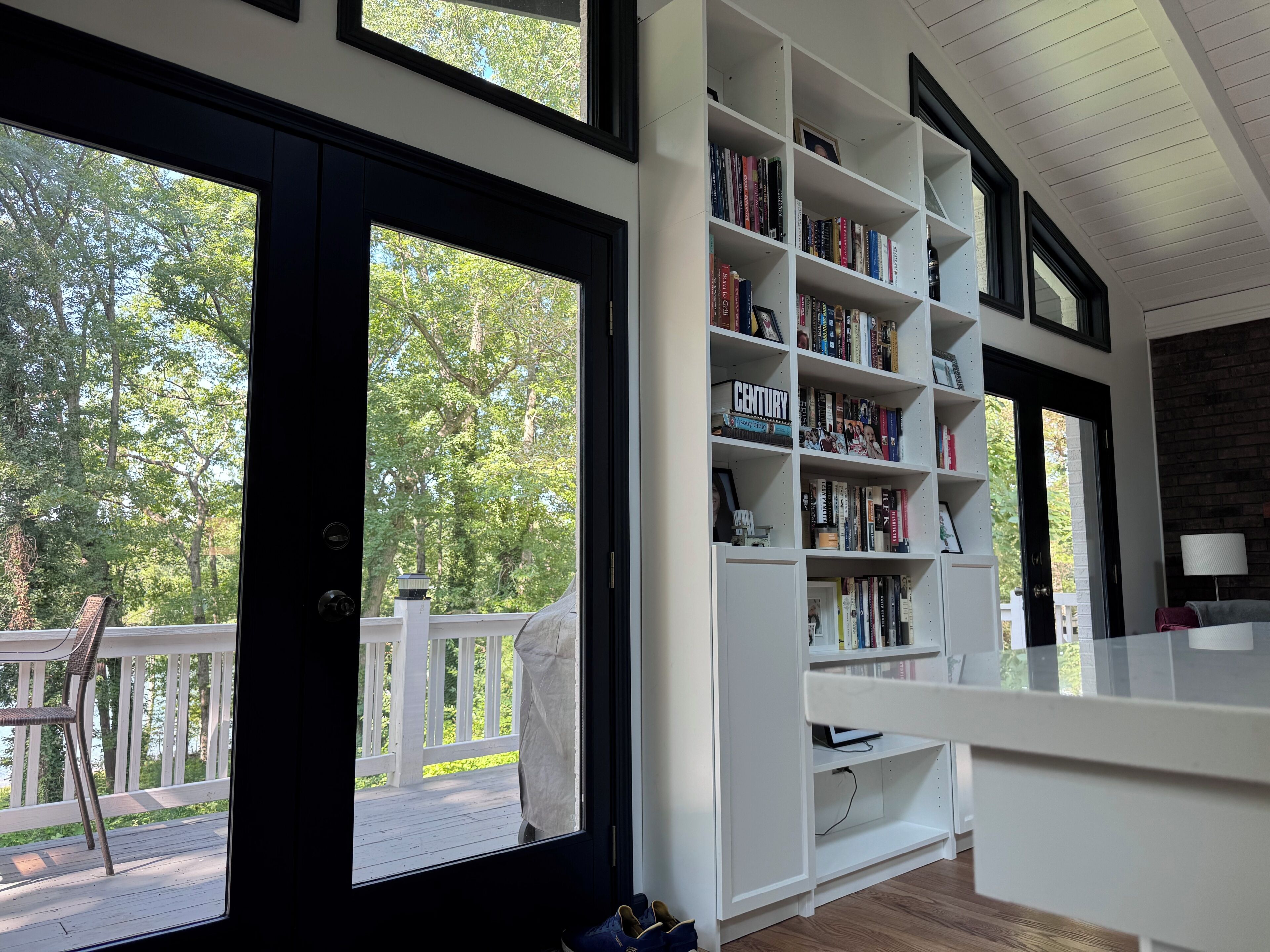 A room with a large bookshelf, a white desk, and a view of a deck with a chair.