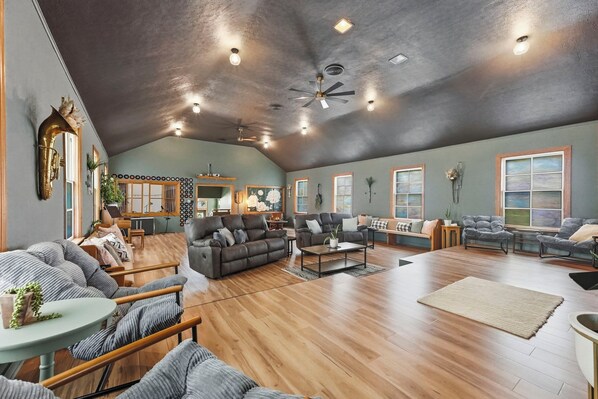 A spacious living room with a high ceiling, multiple seating areas, and a visible kitchen area.