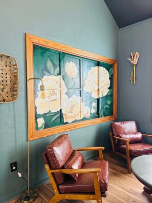 A room with a large floral painting on the wall, two leather chairs, a round table, and a wall-mounted decorative item.