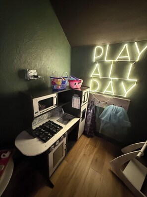 A child's playroom with a kitchenette, a neon 'PLAY ALL DAY' sign, and a blue curtain.