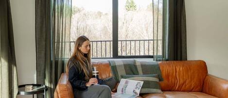 A person sitting on a leather sofa with a magazine, a glass of drink, and a window with curtains in a room with a small table beside the sofa.