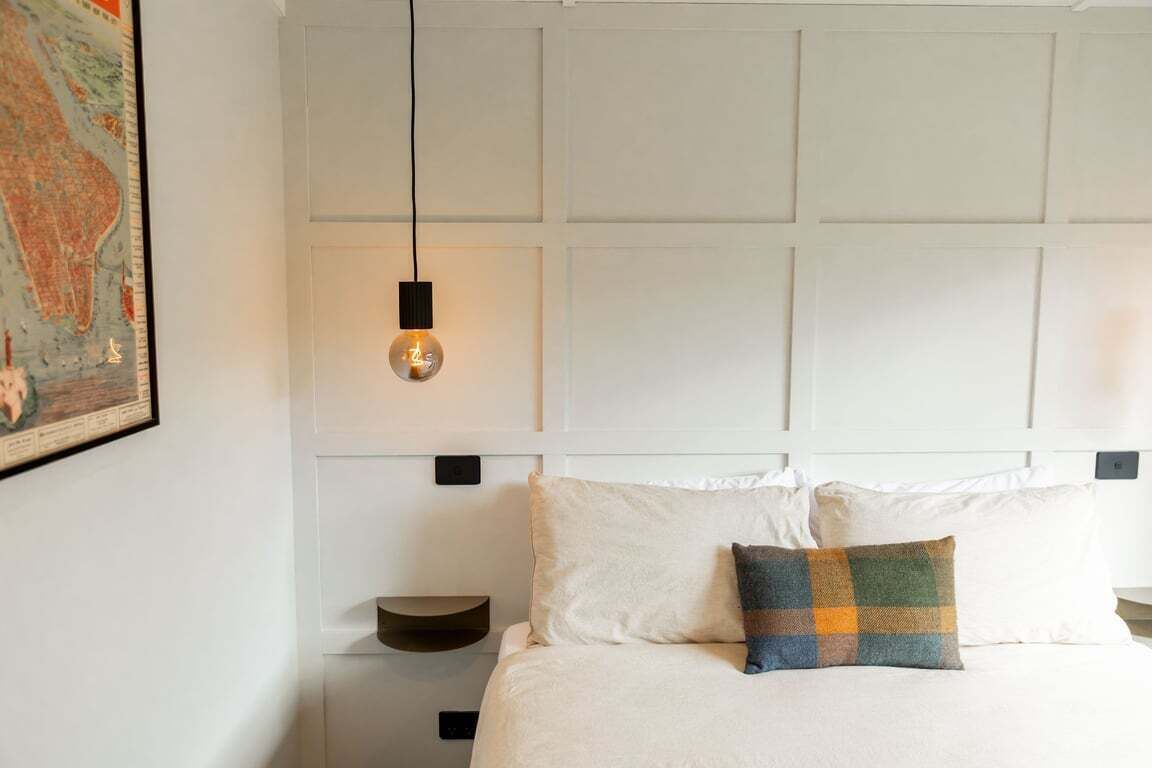 A neatly made bed with a single pillow, a hanging light fixture, and a wall-mounted shelf.