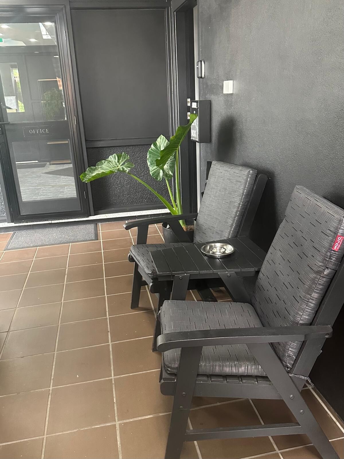 A seating area with two chairs and a table, a potted plant, and a view through a glass door labeled 'OFFICE'.