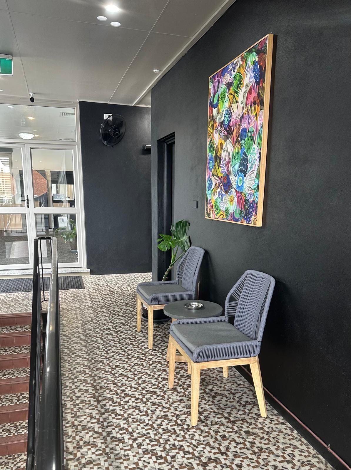 Et receptionsområde med moderne design, med en sort væg, et farverigt abstrakt maleri, to grå polstrede stole, et lille rundt bord og et mønstret gulv.