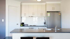 A modern kitchen with white cabinets, a stainless steel refrigerator, a built-in oven, and a bar area with two stools. - Morrison Shorehouse, Middleton Family Beach House (Middleton)