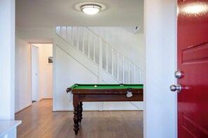 Ein Raum mit Billardtisch, Holzboden, einer Treppe und einer roten Tür.