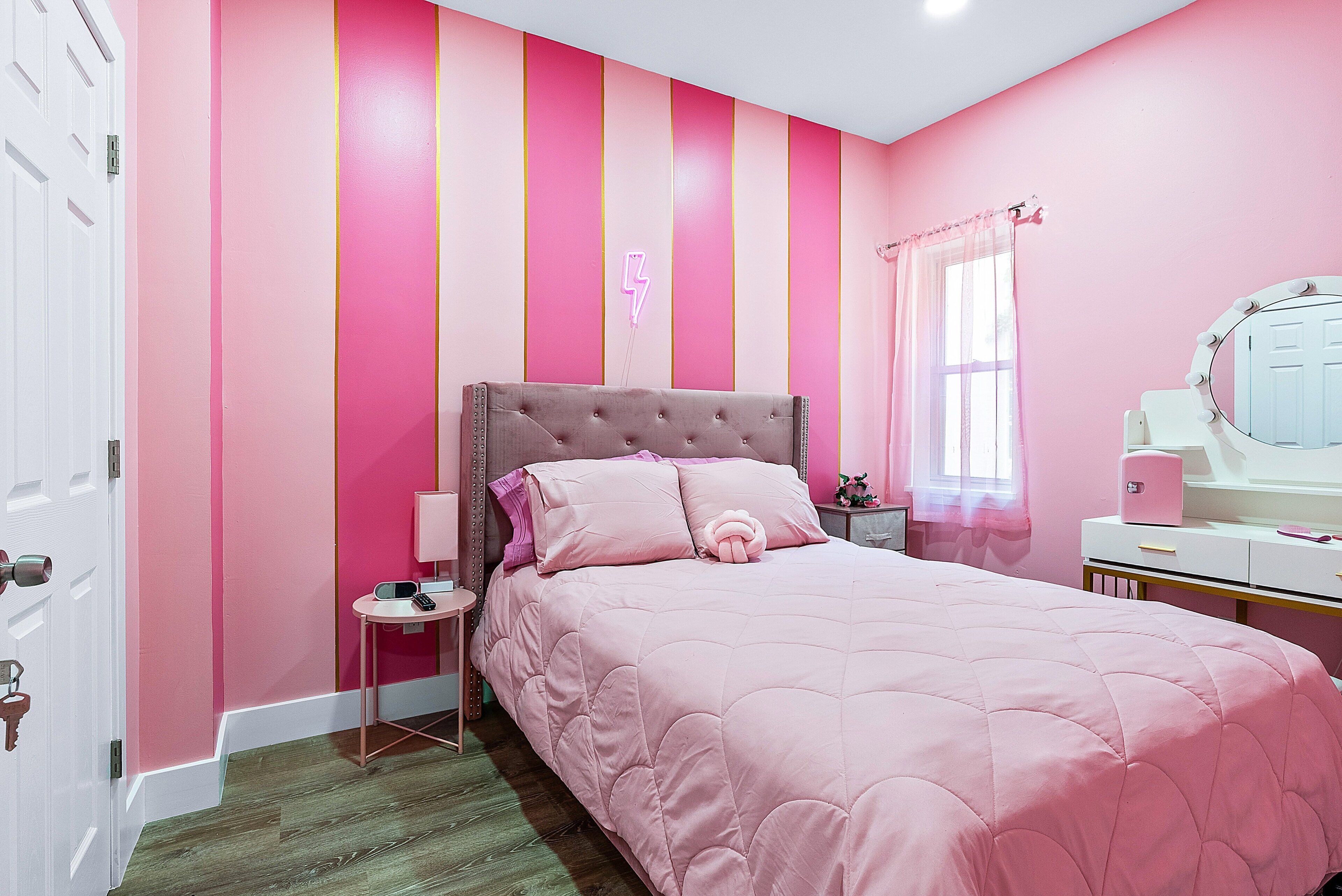 Un dormitorio rosa con cabecera capitonada, mesita de noche con lámpara, espejo y ventana con cortinas translúcidas.
