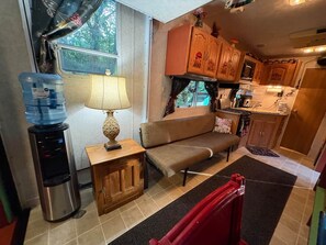 A compact kitchen with a water dispenser, a small table with a lamp, a sofa, and wooden cabinets.