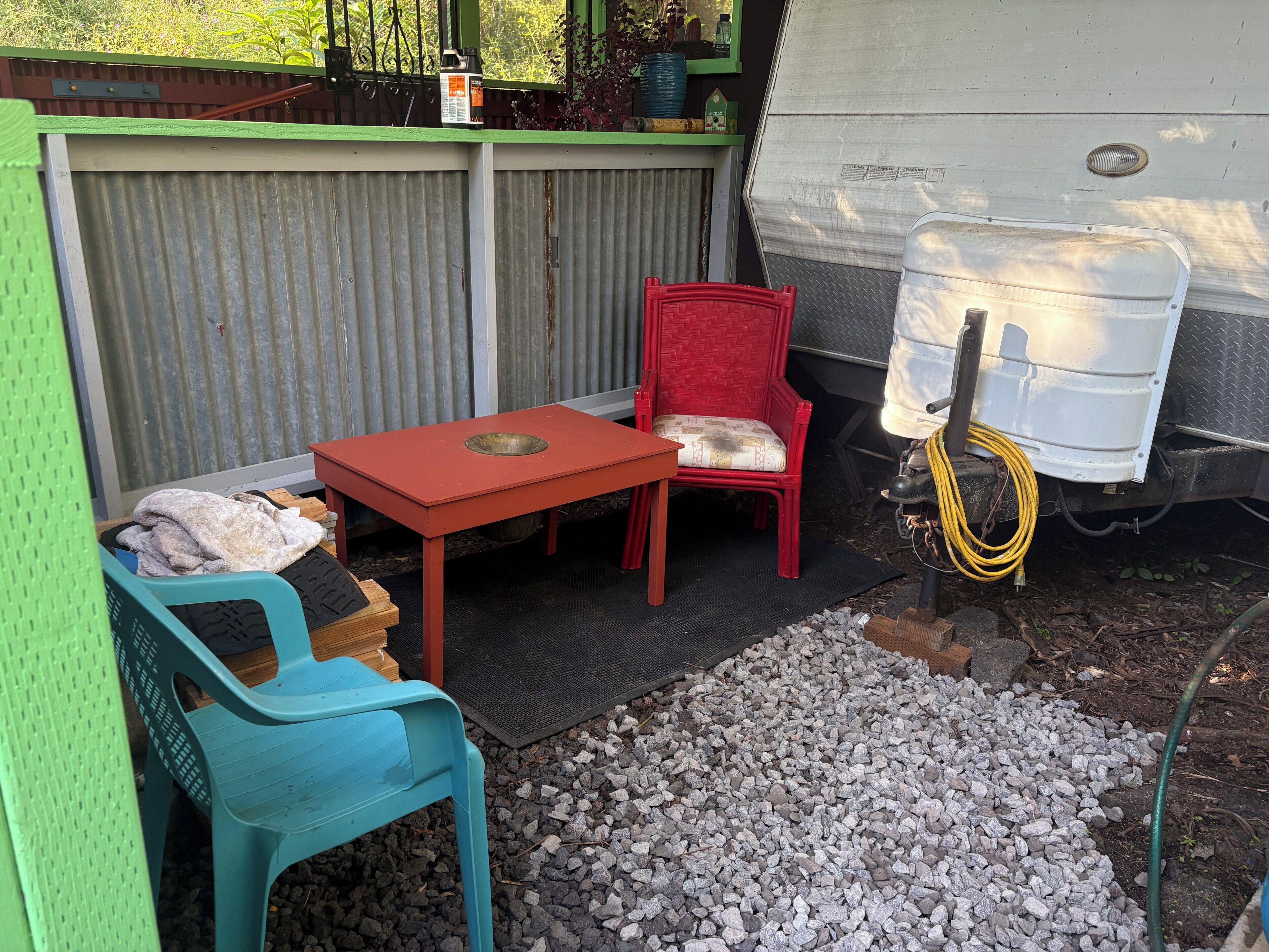 A small outdoor seating area with a table and chairs, a red chair with a cushion, a blue plastic chair, and a white water container.