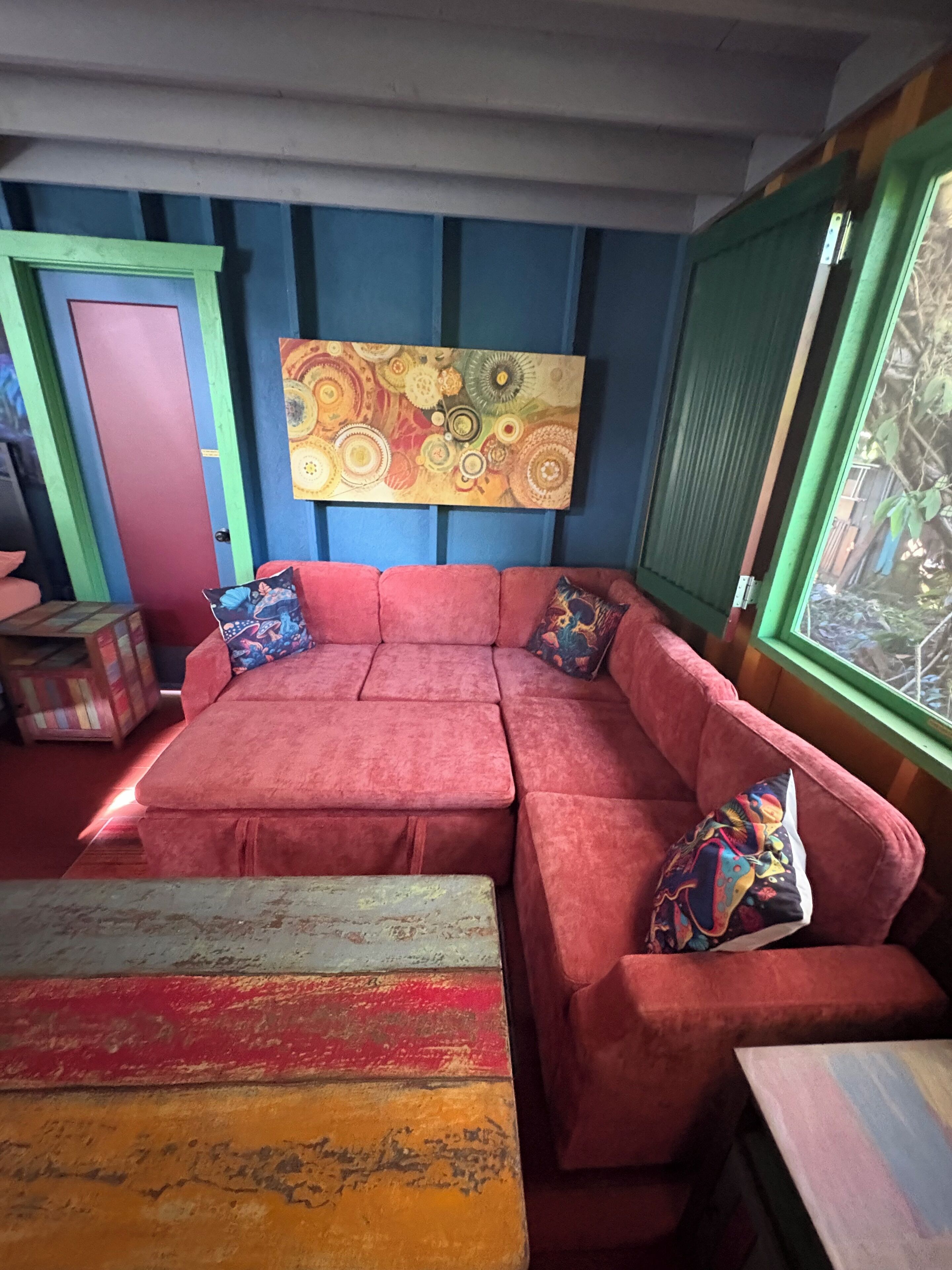 Une pièce avec un canapé rouge, des coussins aux couleurs vives, une table multicolore et un grand tableau abstrait accroché au mur.