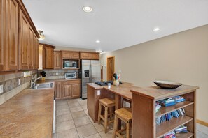 A kitchen with wooden cabinets, a stainless steel refrigerator, a microwave, and a built-in oven. There are two stools at the island and a small shelf with books. - Home with access to the Elk Trail (Angel Fire)