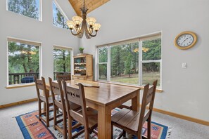 A dining room with a wooden table and chairs, a chandelier, and large windows offering a view of trees. - Home with access to the Elk Trail (Angel Fire)