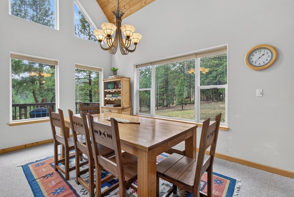 A dining room with a wooden table and chairs, a chandelier, and large windows offering a view of trees. - Home with access to the Elk Trail (Angel Fire)