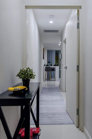 A narrow hallway with a dining area at the end, featuring a table, chairs, and a potted plant.