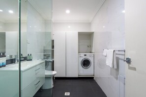 A modern bathroom with a glass shower enclosure, a white sink with toiletries, a washing machine, and a towel rack.