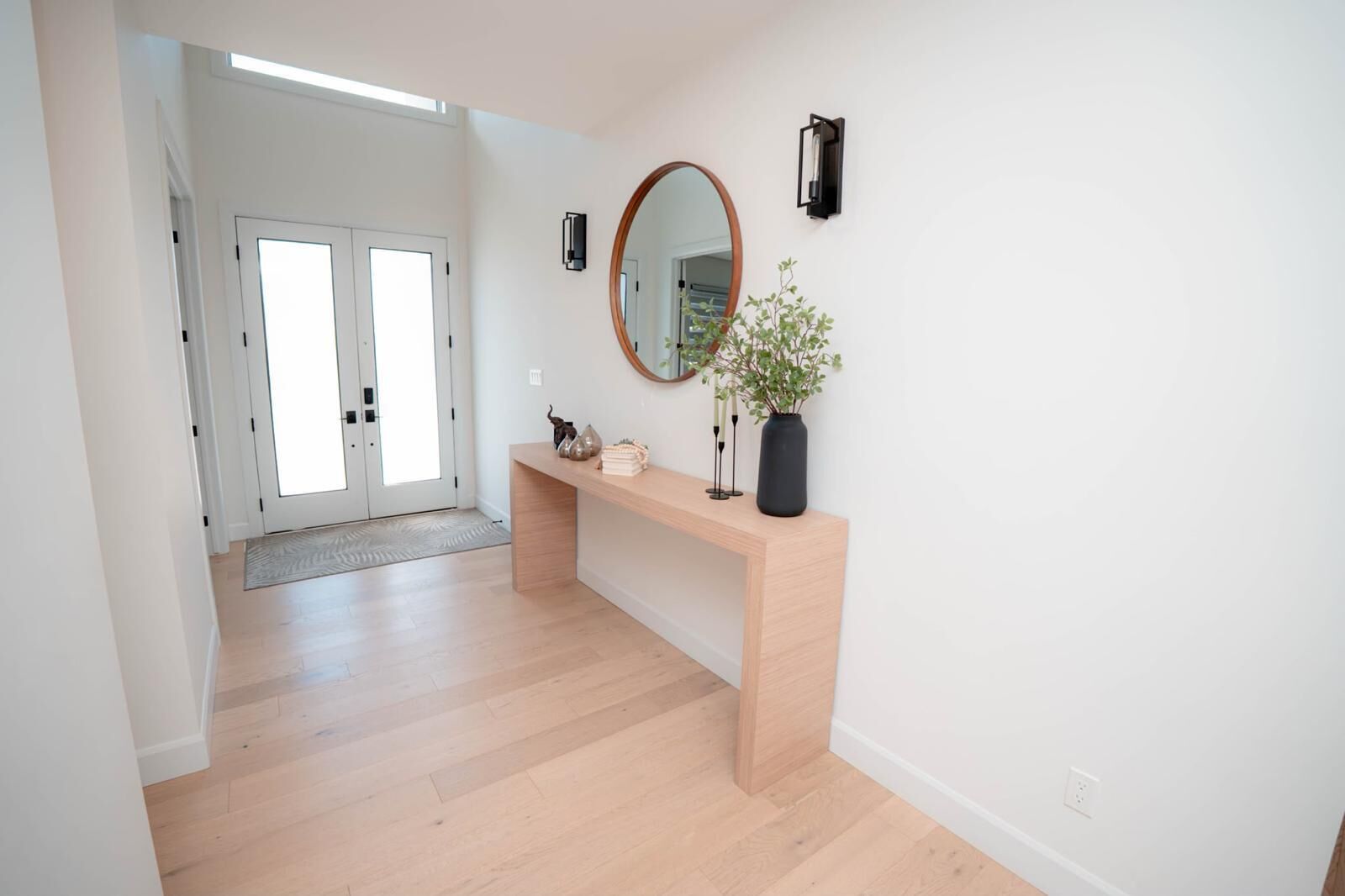 Un couloir doté d’un plancher en bois, d’un banc agrémenté d’objets décoratifs, d’un miroir rond et d’une porte donnant sur une autre pièce.