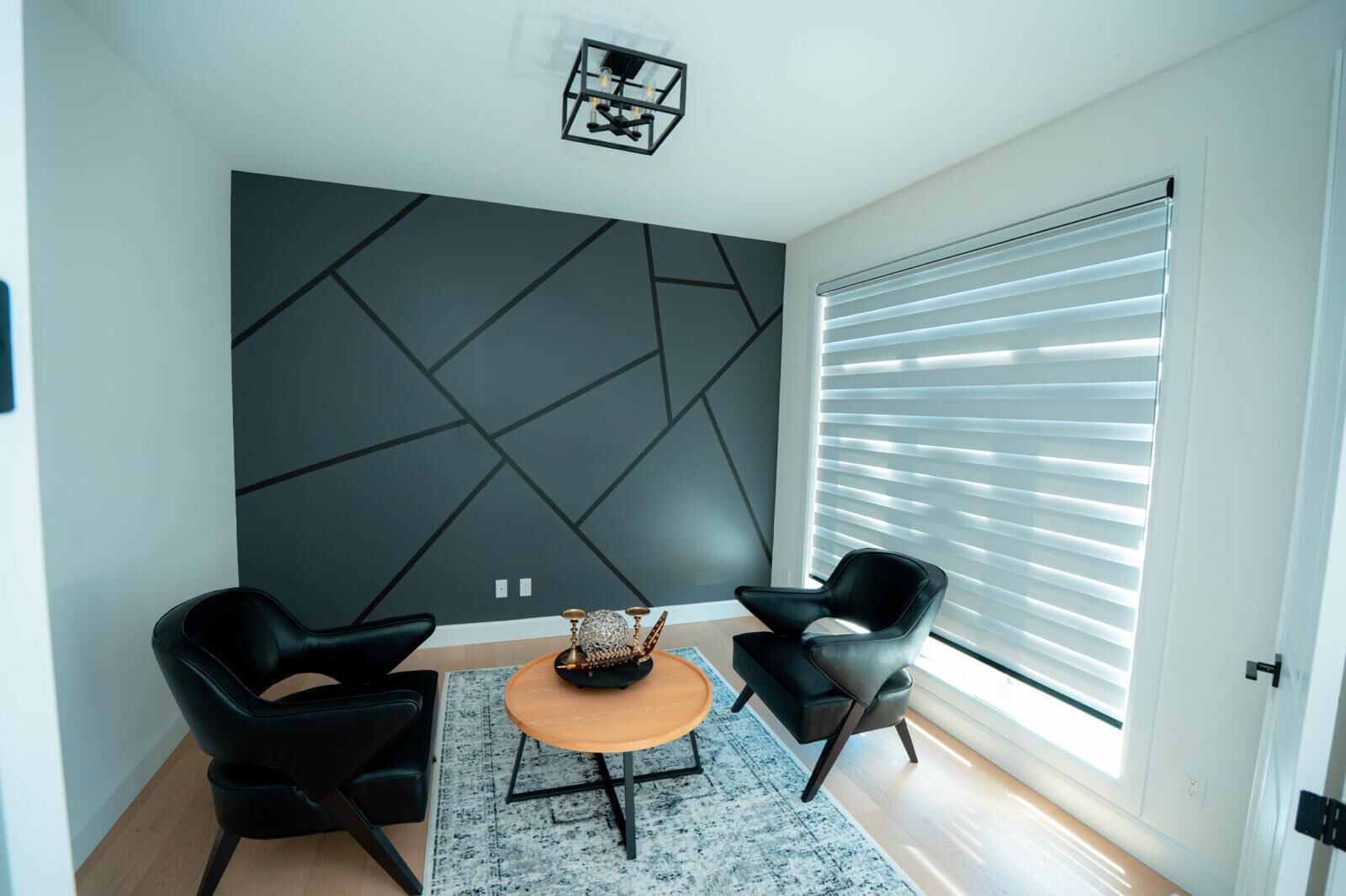 A modern living room with a geometric wall design, two black chairs, a round wooden coffee table, and a large window with blinds.