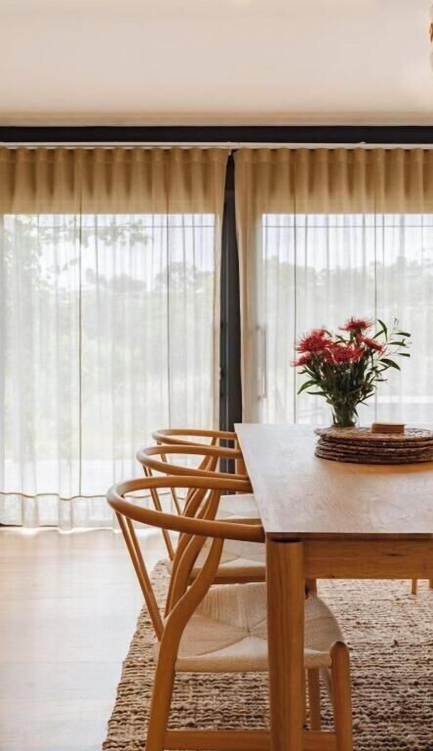 A dining area with a wooden table and chairs, a vase with flowers, and sheer curtains.