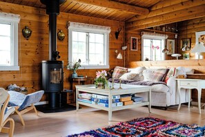 Un acogedor interior de cabaña con estufa de leña, un sofá, una mesa de centro con libros y una alfombra colorida.