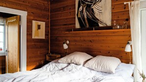 A wooden room with a bed, two bedside lamps, a painting, and a window with curtains. - Traditional cabin among the dunes (Skagen)