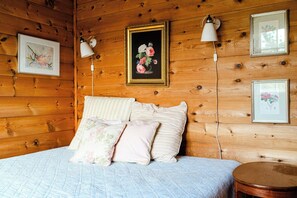 Un dormitorio de cabaña de madera con una cama, almohadas, un cuadro floral enmarco y lámparas fijadas a la pared.