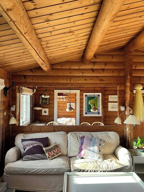 Interior acogedor de una cabaña con techo de madera, un sofá con cojines estampados, una zona de comedor con sillas y una mesa con lámpara.