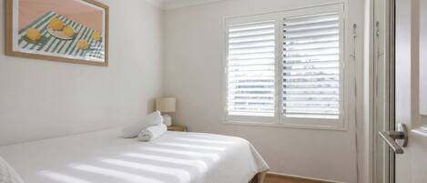 A bedroom with a bed, a bedside lamp, a window with blinds, and a ceiling fan.