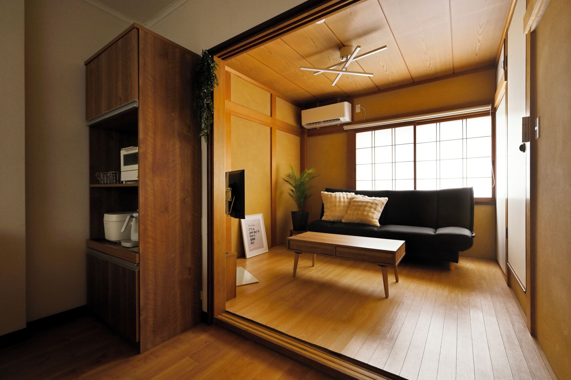 A room with a wooden floor, a black sofa with cushions, a small table, a potted plant, and a ceiling fan.