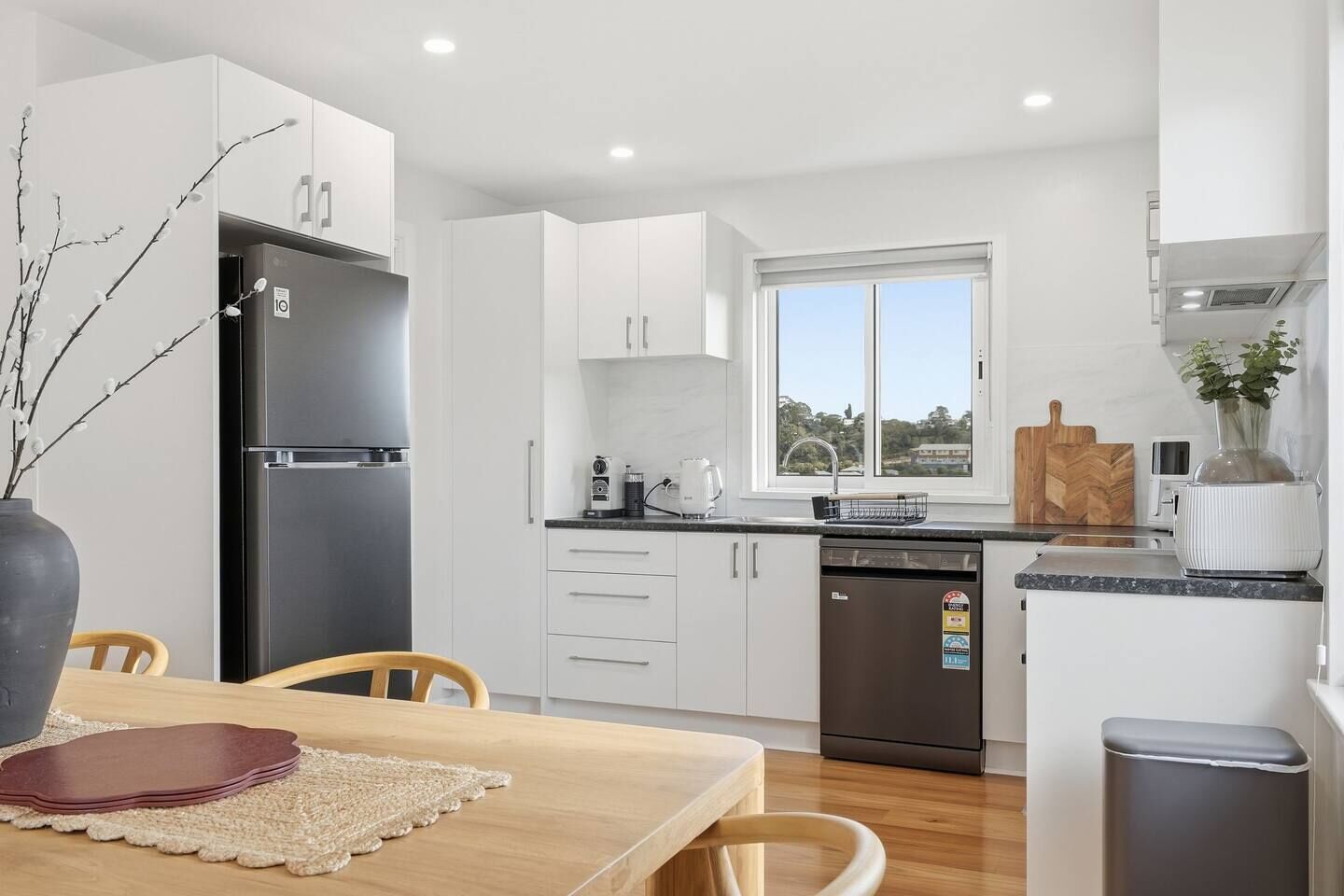 A modern kitchen with white cabinets, a black refrigerator, a dishwasher, and a window with a view.
