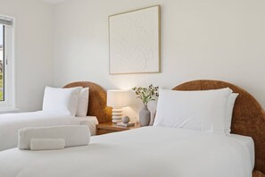 A hotel room with two beds, a nightstand with a lamp, a vase with flowers, and a framed artwork on the wall. - Modern 3BR Home Near Bellerive (Warrane)